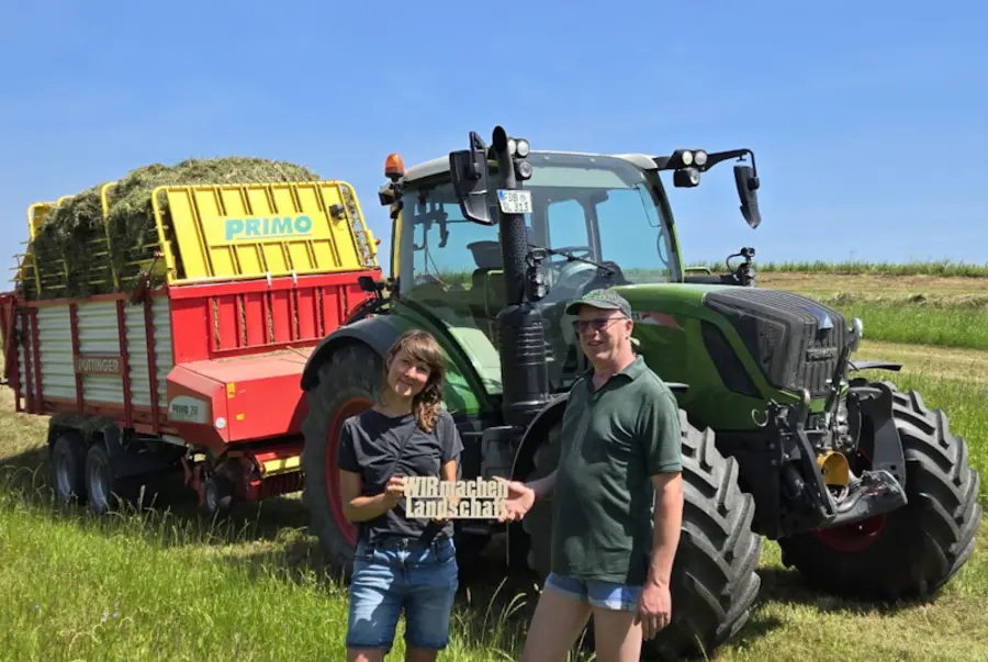 Landwirt und Landschaftspflegerin vor einem Traktor mit Anhänger.