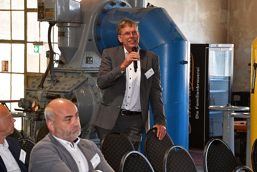 ©swa/Thomas Hosemann Mann im grauen Anzug spricht mit einem Mikrofon vor einer großen Industriemaschine