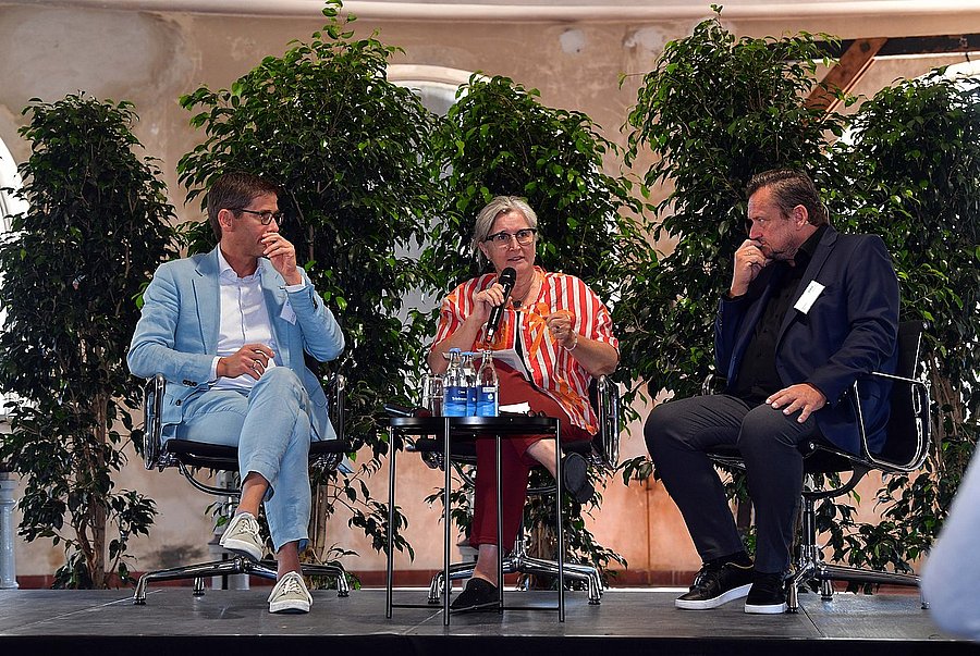 ©swa/Thomas Hosemann Frau spricht auf einem Podium ins Mikrofon, während zwei Männer neben ihr sitzen und zuhören