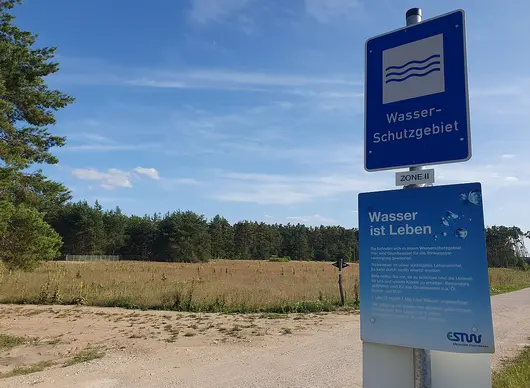 Schild „Wasserschutzgebiet“ an einer offenen Wiesenfläche vor einem Wald unter blauem Himmel